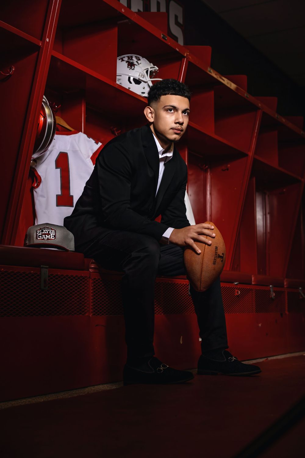A person in a black suit sits in a red-toned locker room holding a football while wearing formal attire.