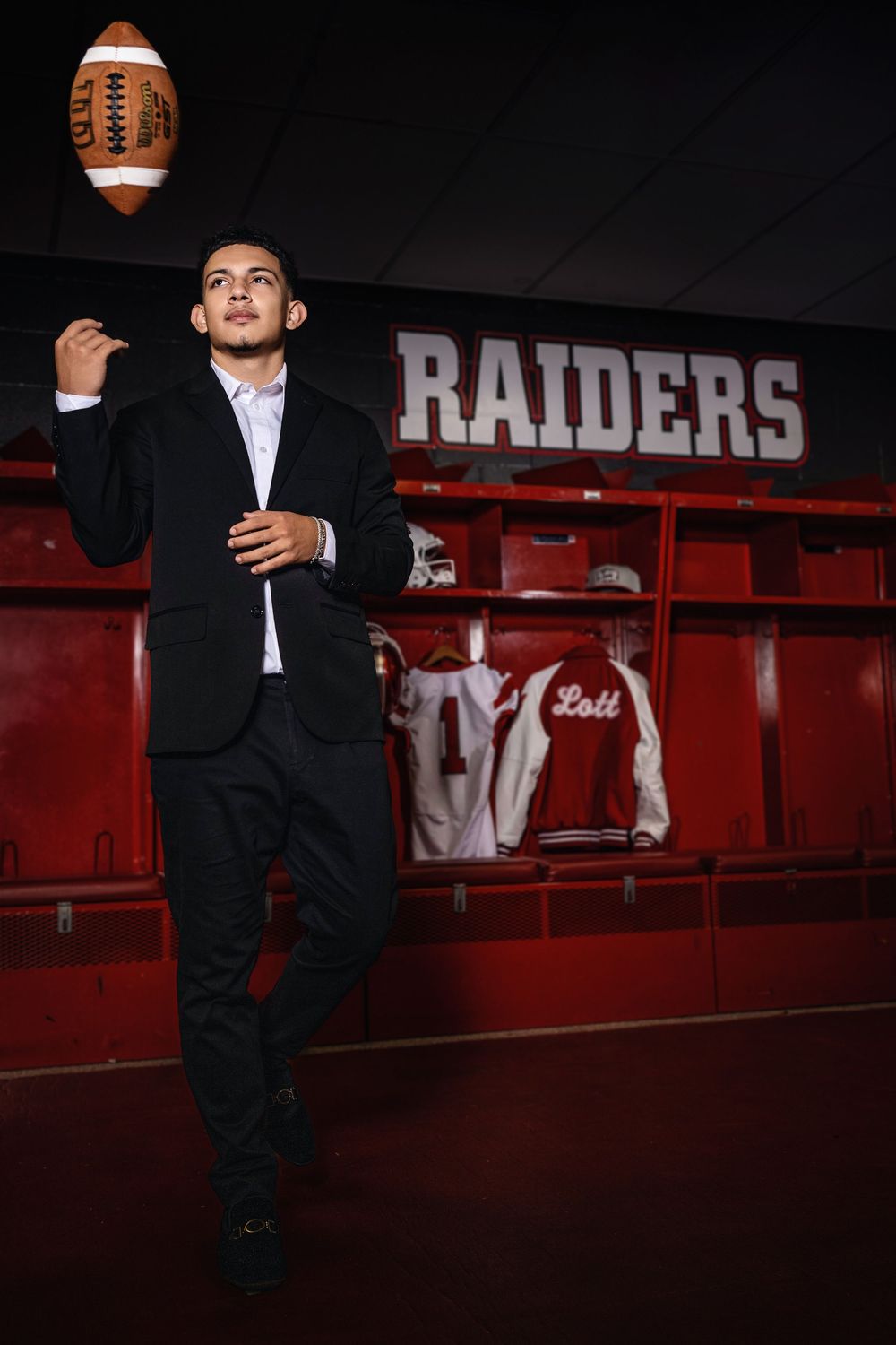 A person in a suit tosses a football in front of a Raiders team locker room sign and red lockers.