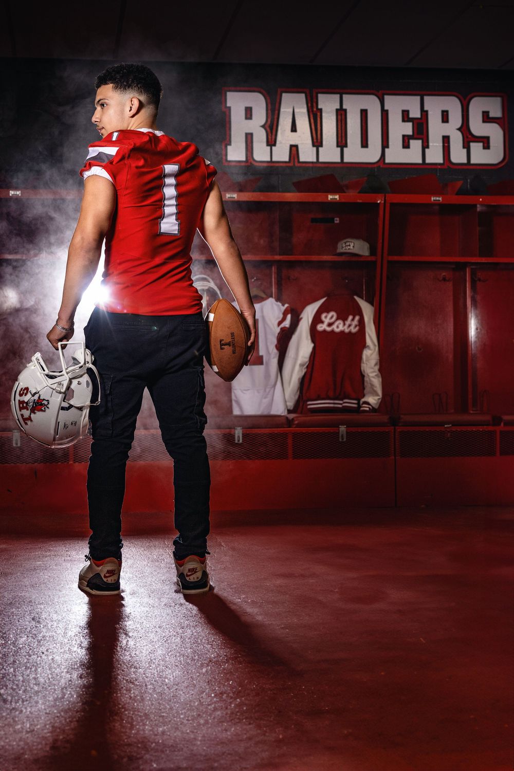 Football player in red Raiders jersey stands in locker room holding helmet and football in dramatic lighting.