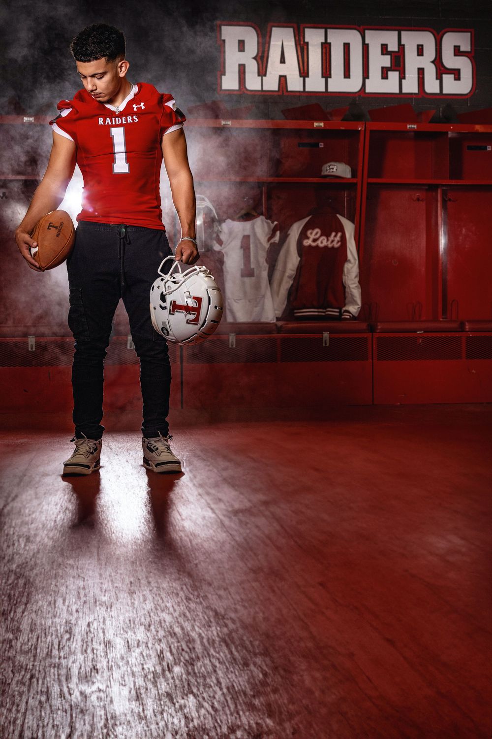 Football player stands in red Raiders uniform holding helmet and ball in dramatic locker room lighting effects.