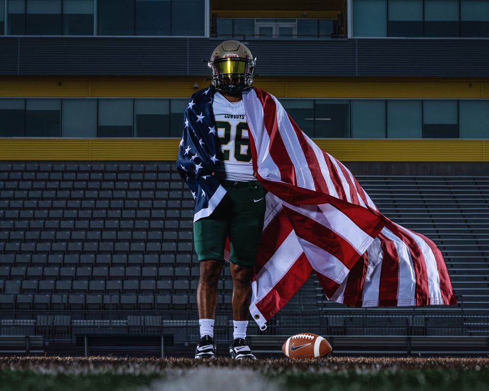 American flag waves during a high school football game with stadium lights illuminating the field at dusk.
