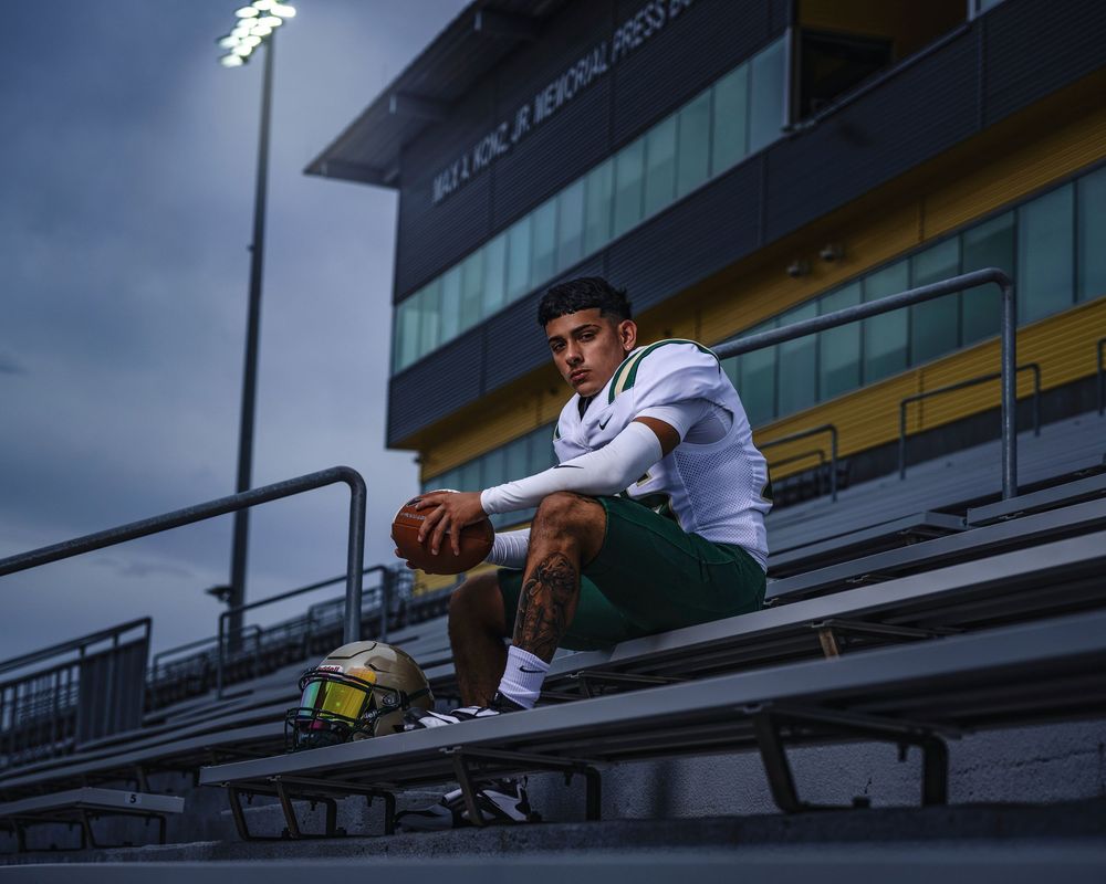 An athlete in uniform poses on stadium bleachers under dramatic cloudy skies.