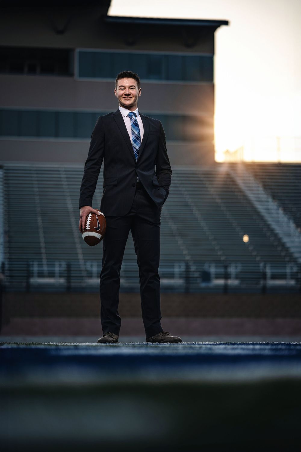 An athletic figure in a dark business suit holds a football while standing in an empty stadium at sunset.