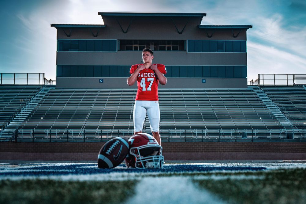 A football player in red jersey number 27 stands on field with helmet on ground and empty stadium bleachers behind.