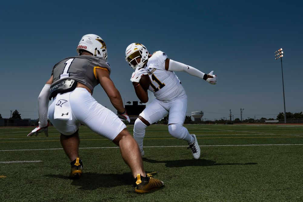 Two football players in white uniforms practice blocking drills on a sunny day during training camp.