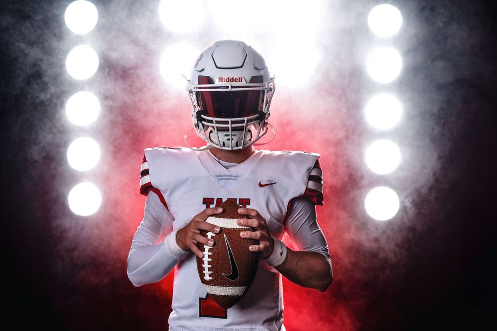 Football player in white uniform holds ball against dramatic red-lit backdrop with glowing spotlights creating atmospheric haze.
