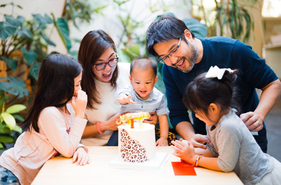 A family celebrates around a birthday cake at a table in a sunlit room with plants in the background.