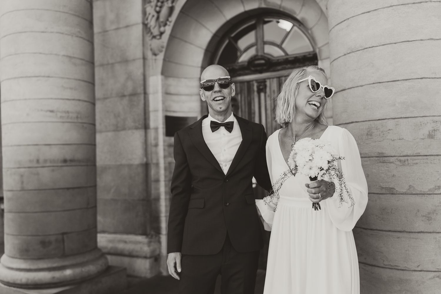 A stylish black and white photo of a bride and groom wearing sunglasses outside a classical building with columns.