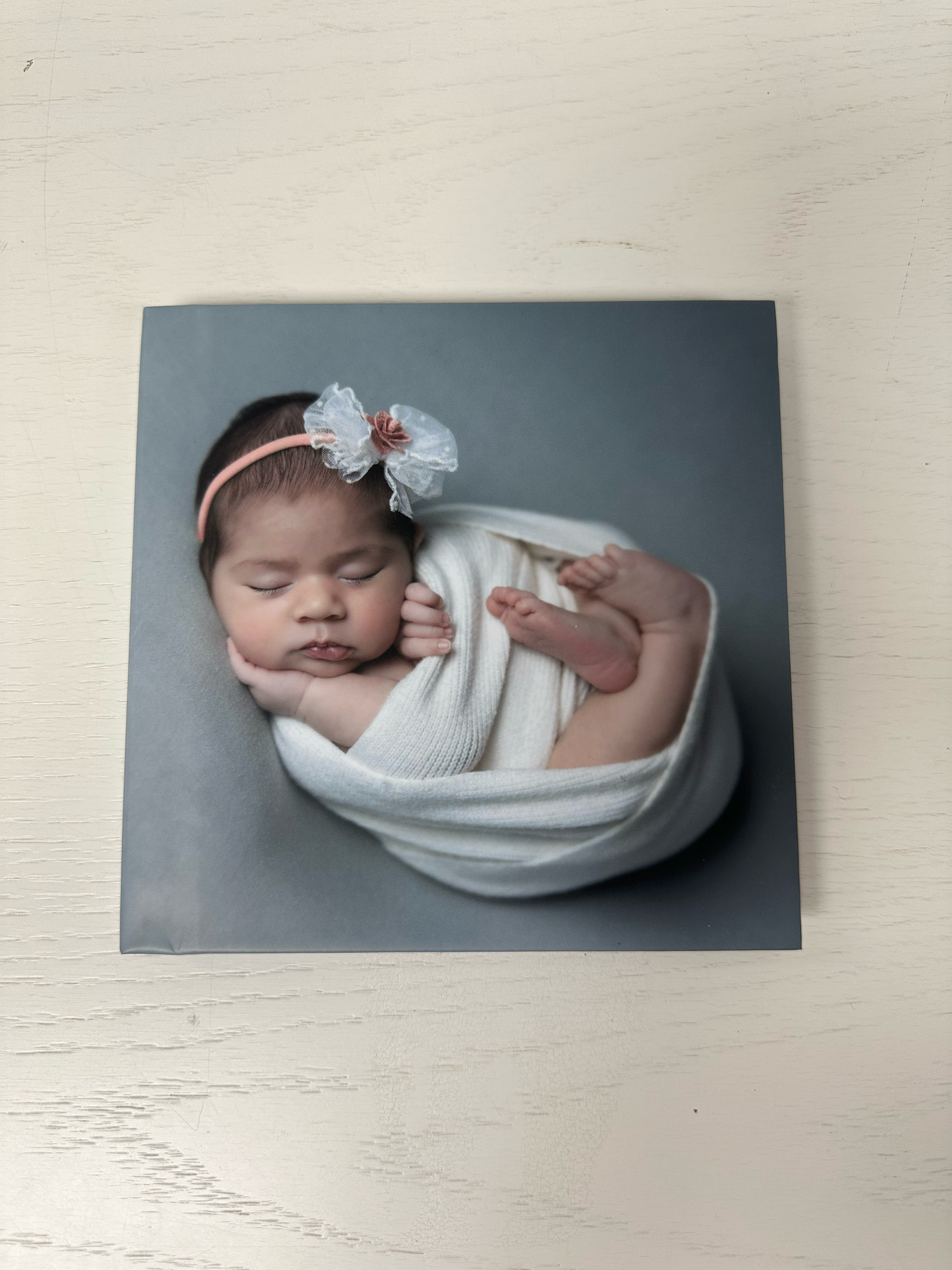 Newborn baby girl sleeping peacefully wrapped in white knit swaddle with floral headband on gray background. Photo book
