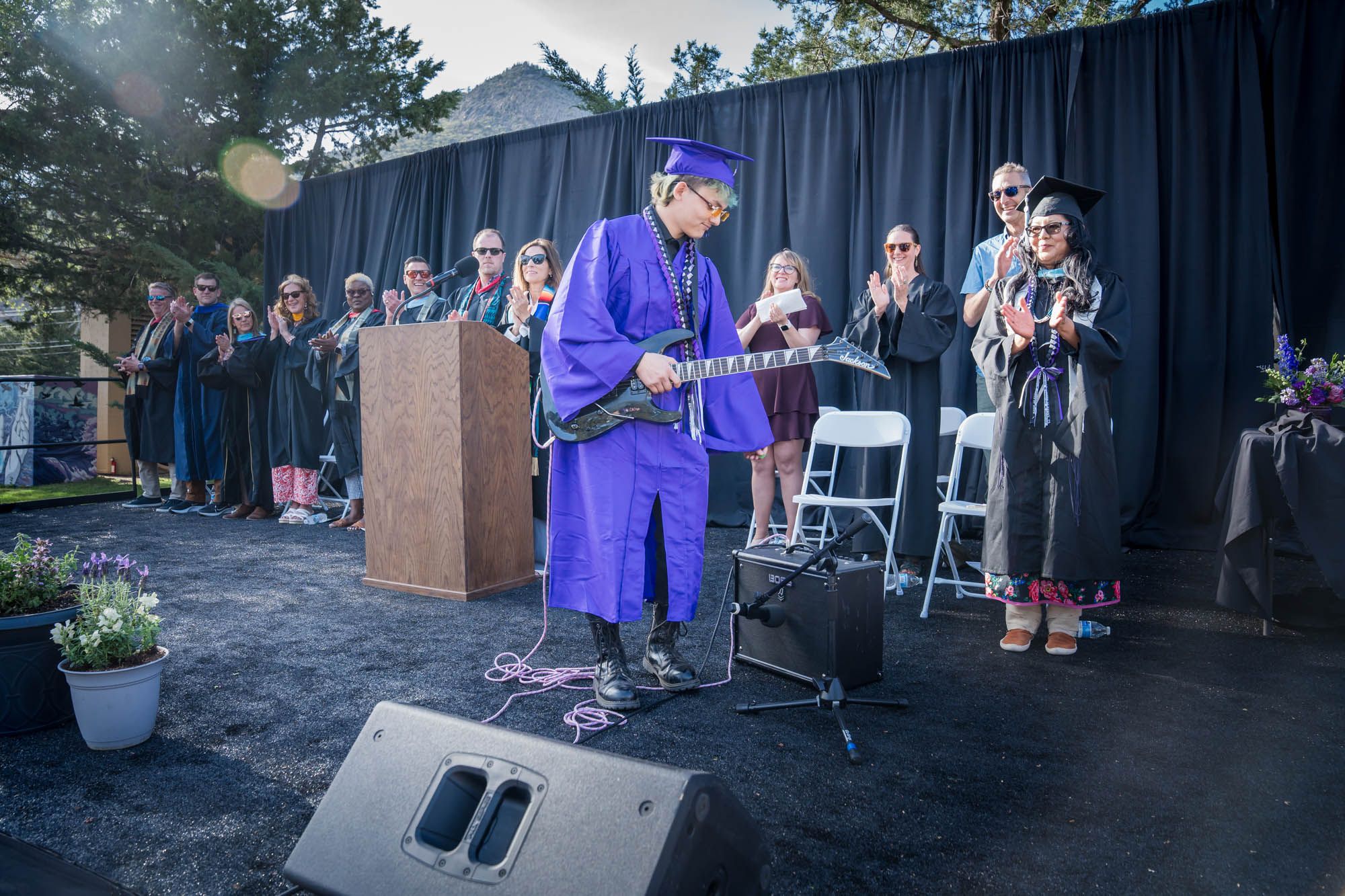 Graduate in purple cap and gown stands at podium during outdoor commencement ceremony with audience in background.