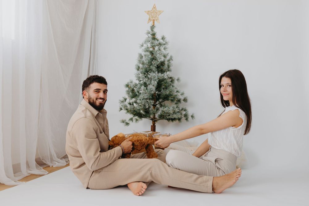 A romantic couple sits cross-legged in front of a Christmas tree wearing neutral-toned outfits.