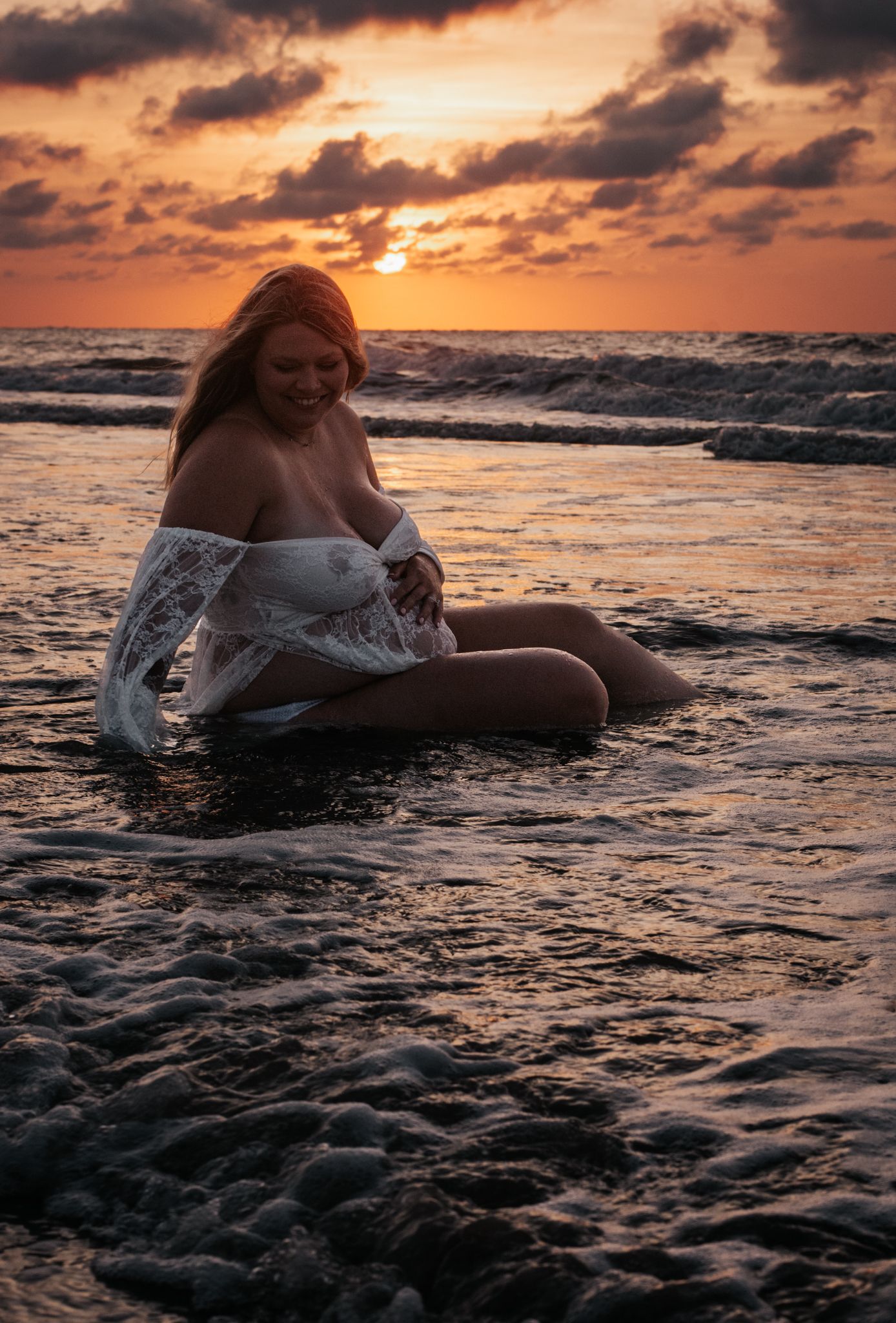 Woman in white dress sitting in shallow ocean water at golden hour sunset.