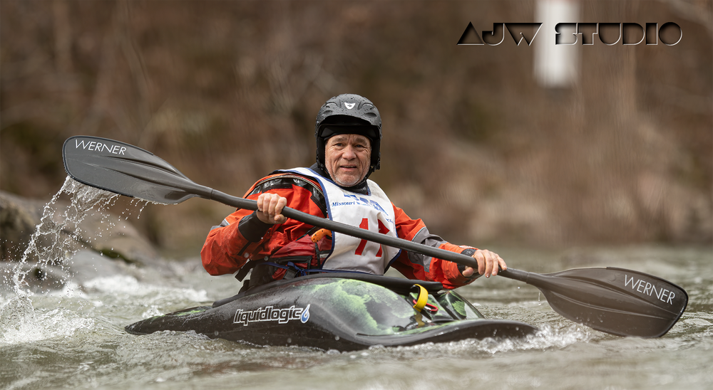 Kayaker in red safety gear paddles through churning whitewater during a competitive event.