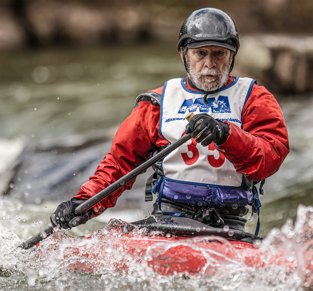 Competitive kayaker in red jersey and black helmet paddles through rough waters during race.