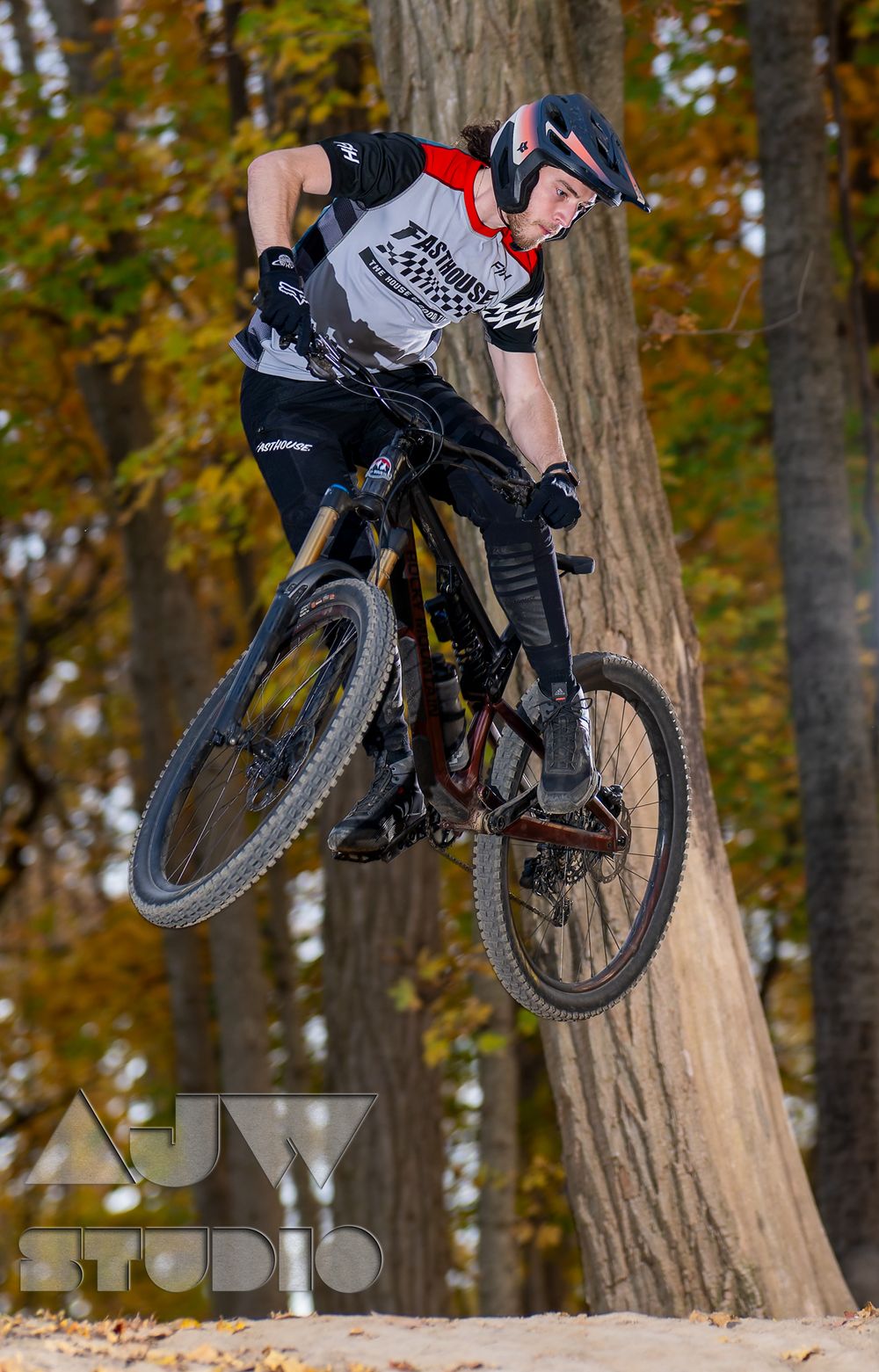 Mountain biker performs a jump on a dirt trail surrounded by autumn trees.