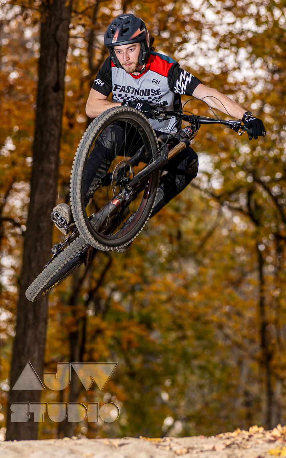 Mountain bike rider executes aerial trick off dirt jump in fall forest setting.