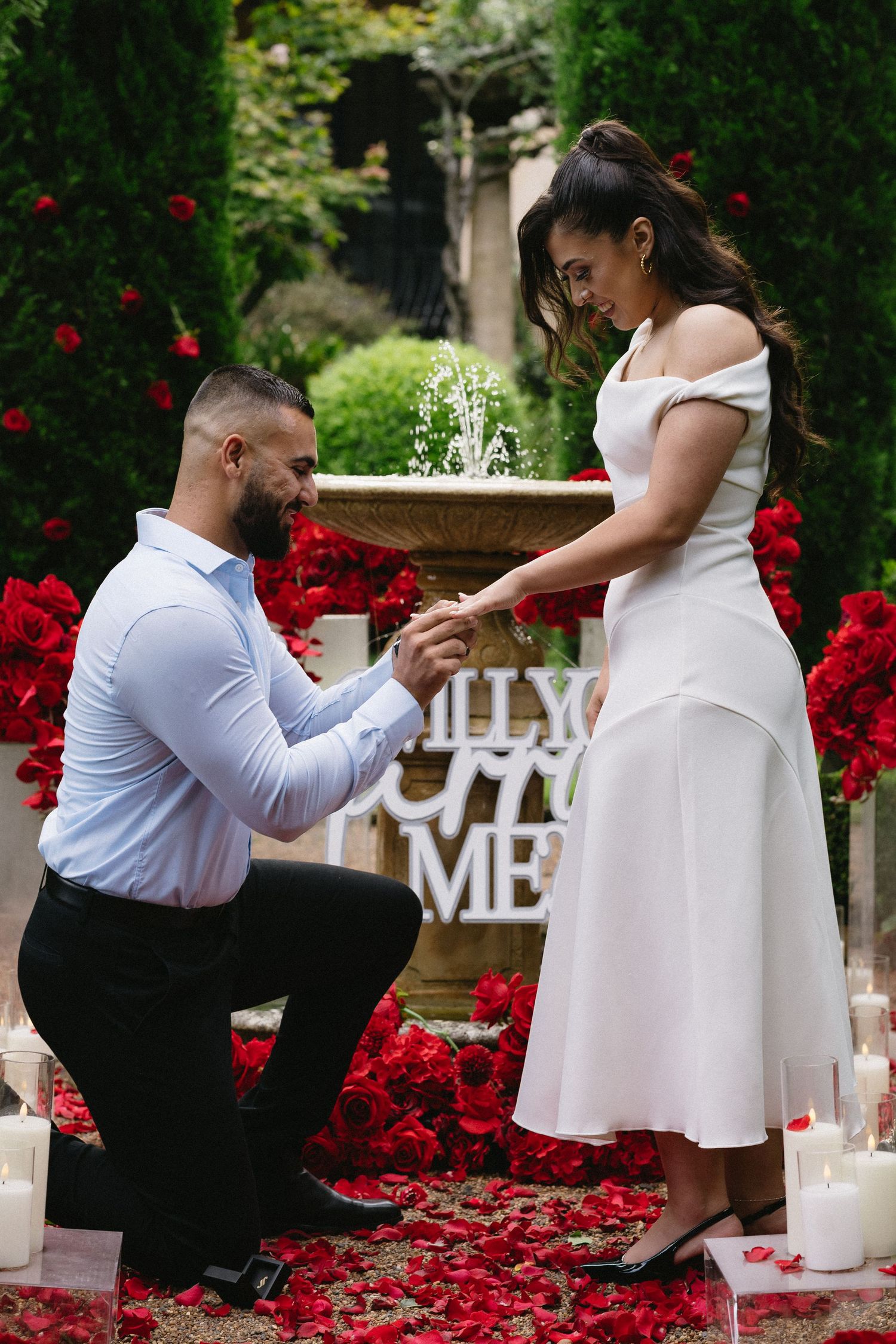Romantic proposal scene with man kneeling before woman among red roses and candles.