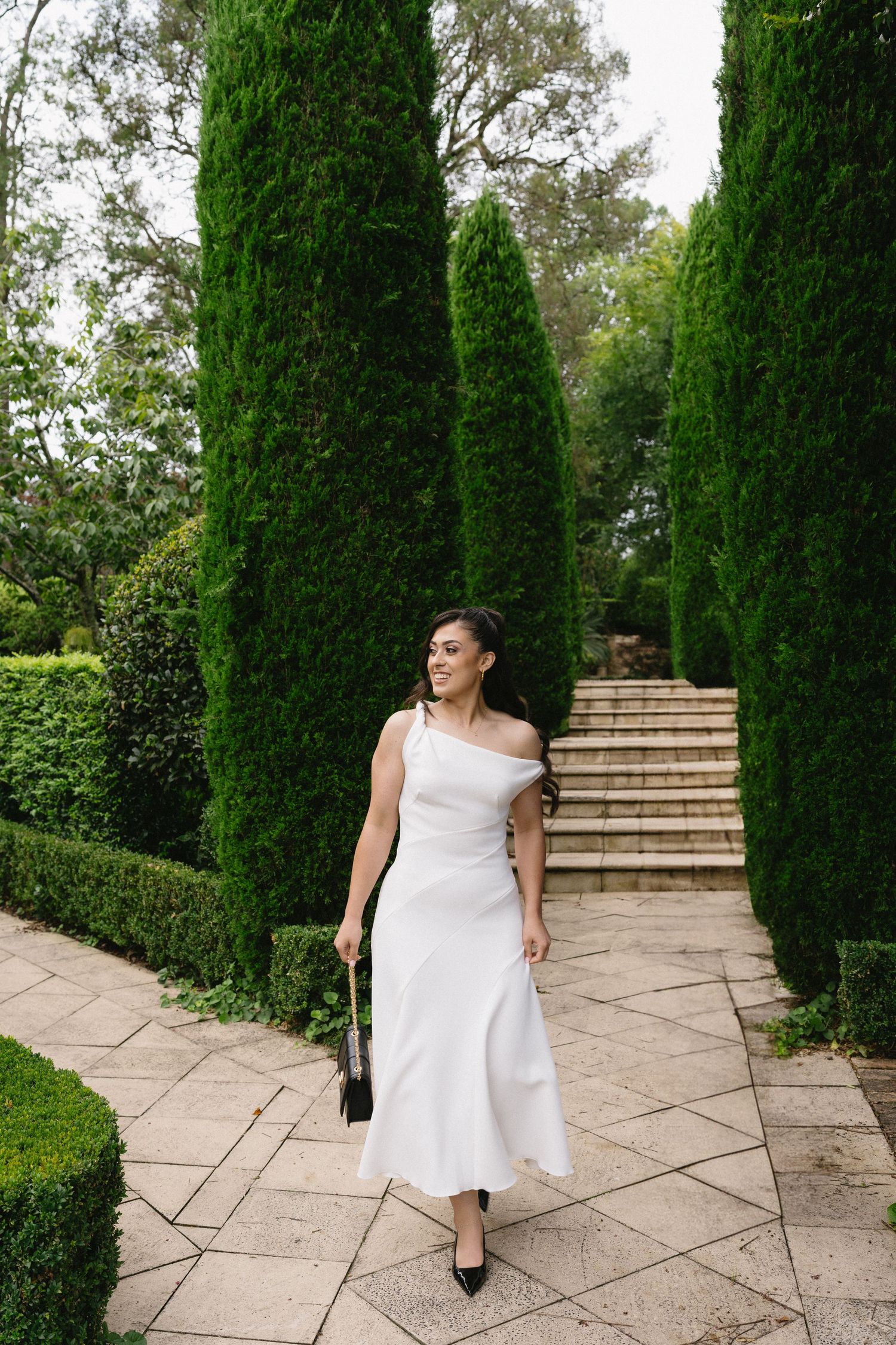 Woman in white dress walking along stone path lined with tall cypress trees.