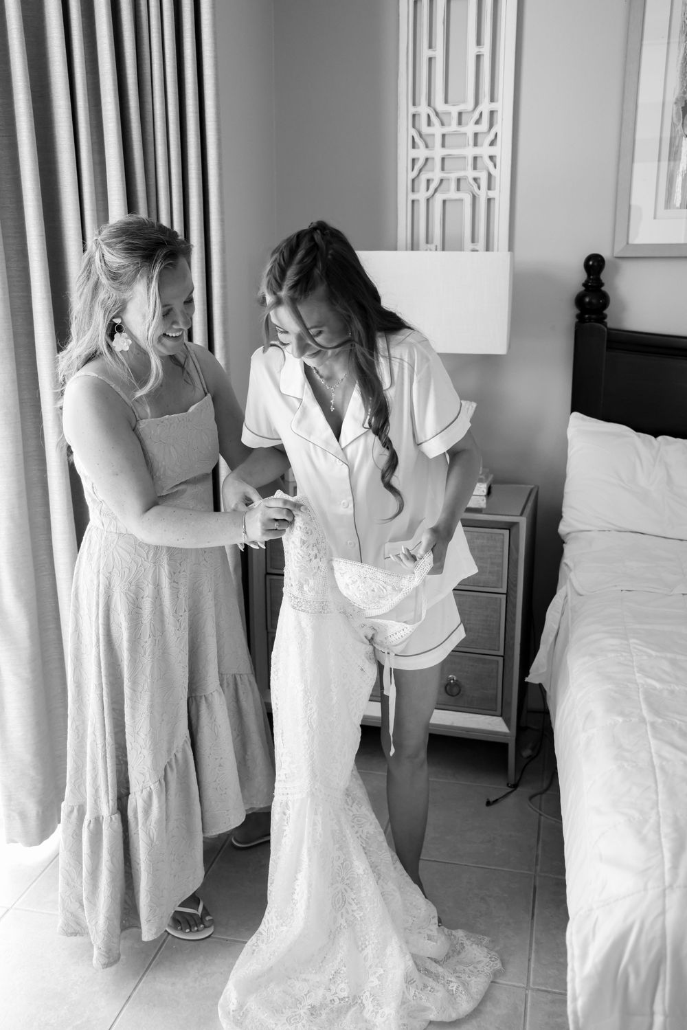 Two people in light colored dresses help with wedding preparations in a bedroom setting.