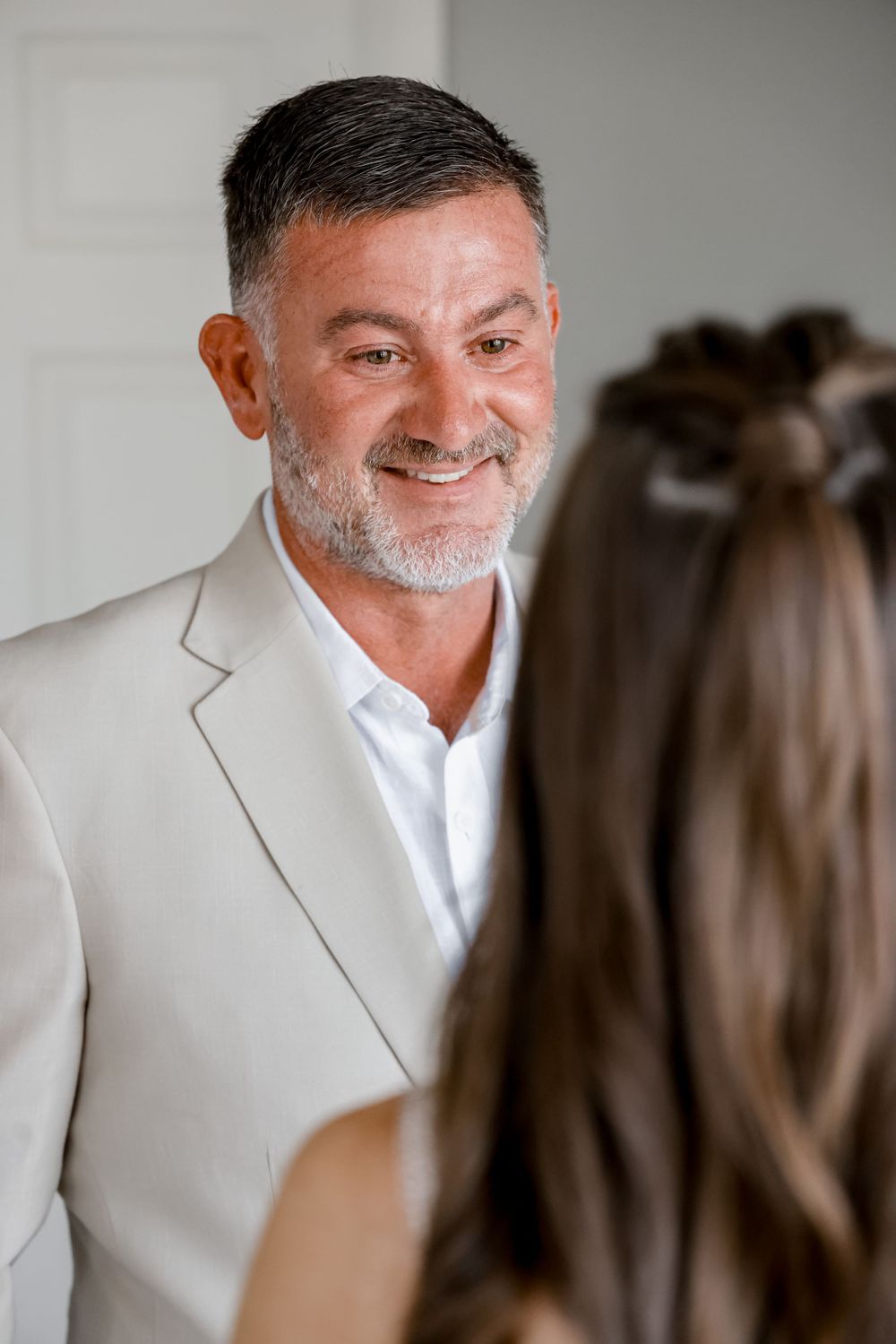 A person wearing a light gray suit jacket smiles warmly in an indoor setting.