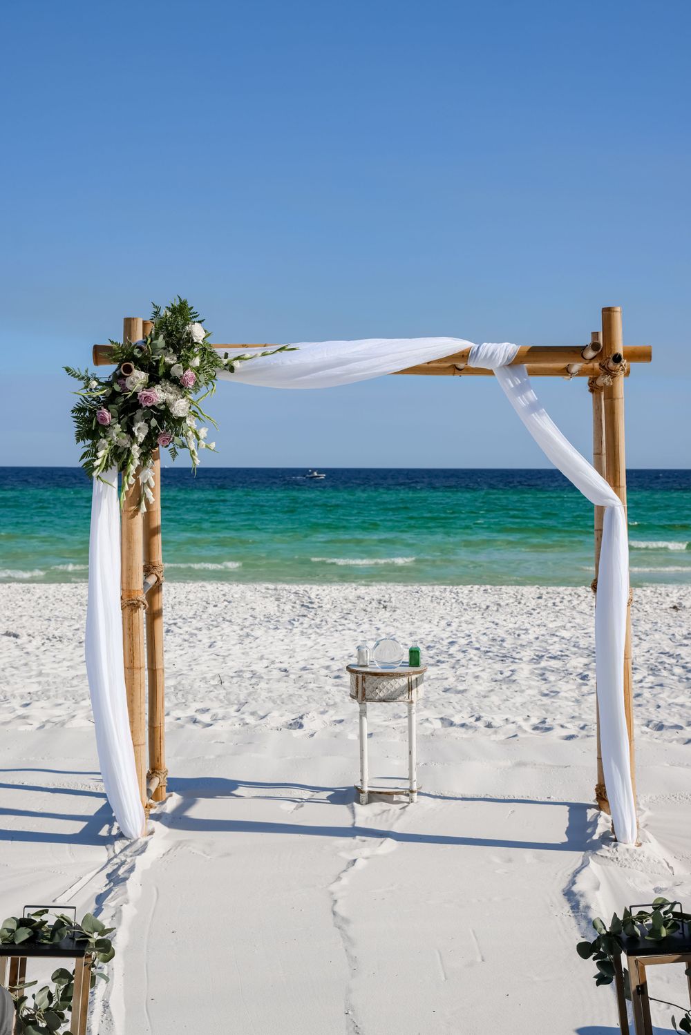 Beach wedding arch with white fabric and floral decorations against turquoise ocean backdrop.