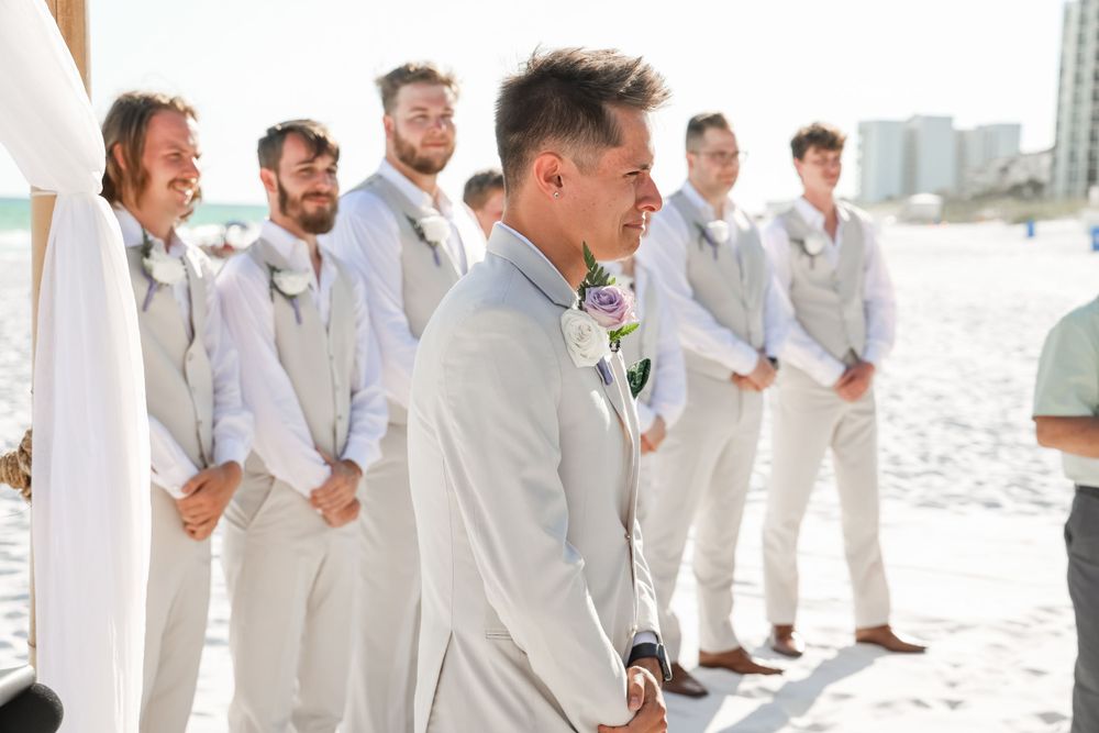 Wedding party in light gray suits standing on white sand beach under bright blue sky.