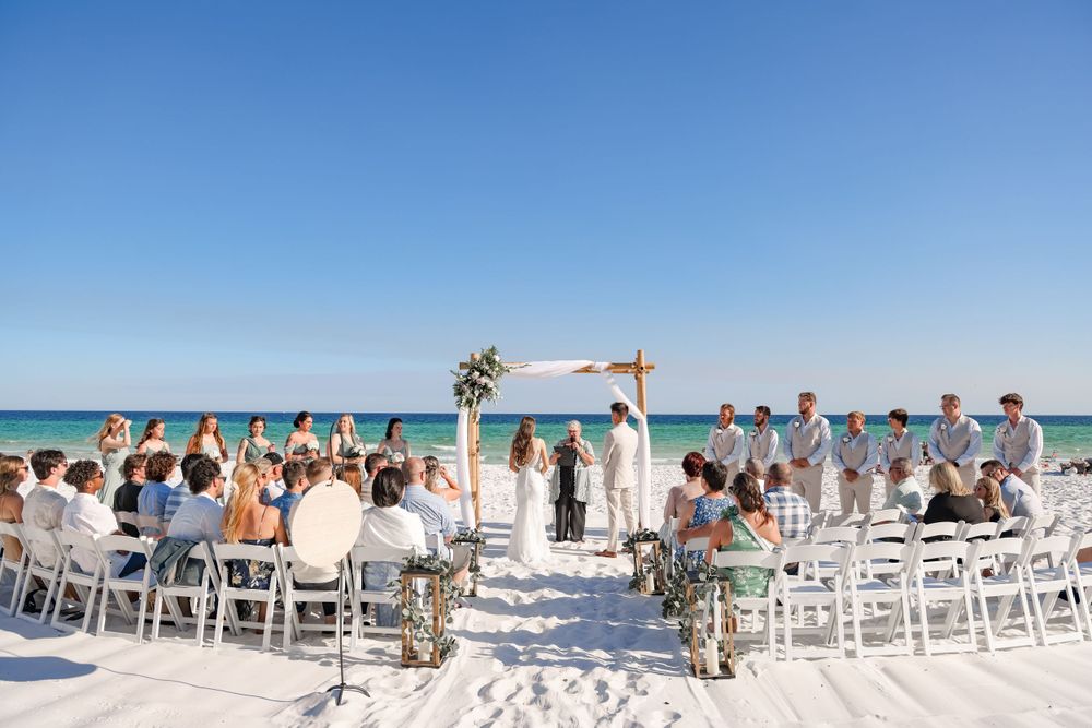 Wide view of beach wedding ceremony setup with white chairs and bamboo arch against ocean backdrop.