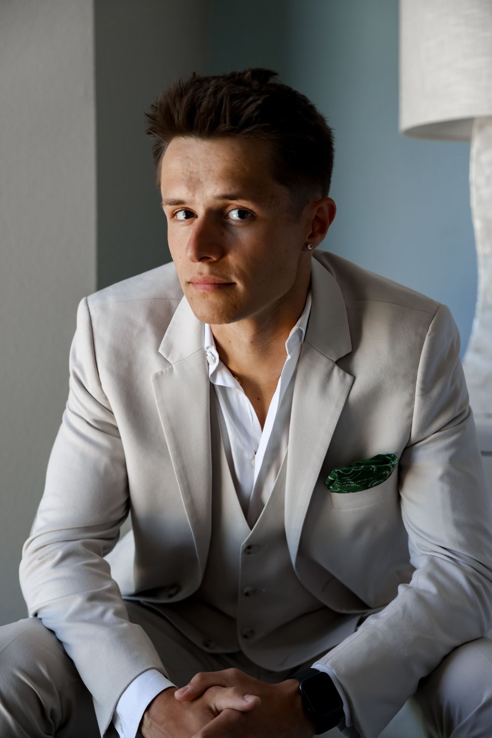 A man in a light gray suit and white shirt sits in an elegant pose against a neutral background.