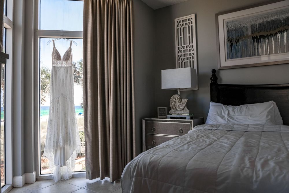 A white wedding dress hangs by a window in a hotel bedroom with neutral decor.
