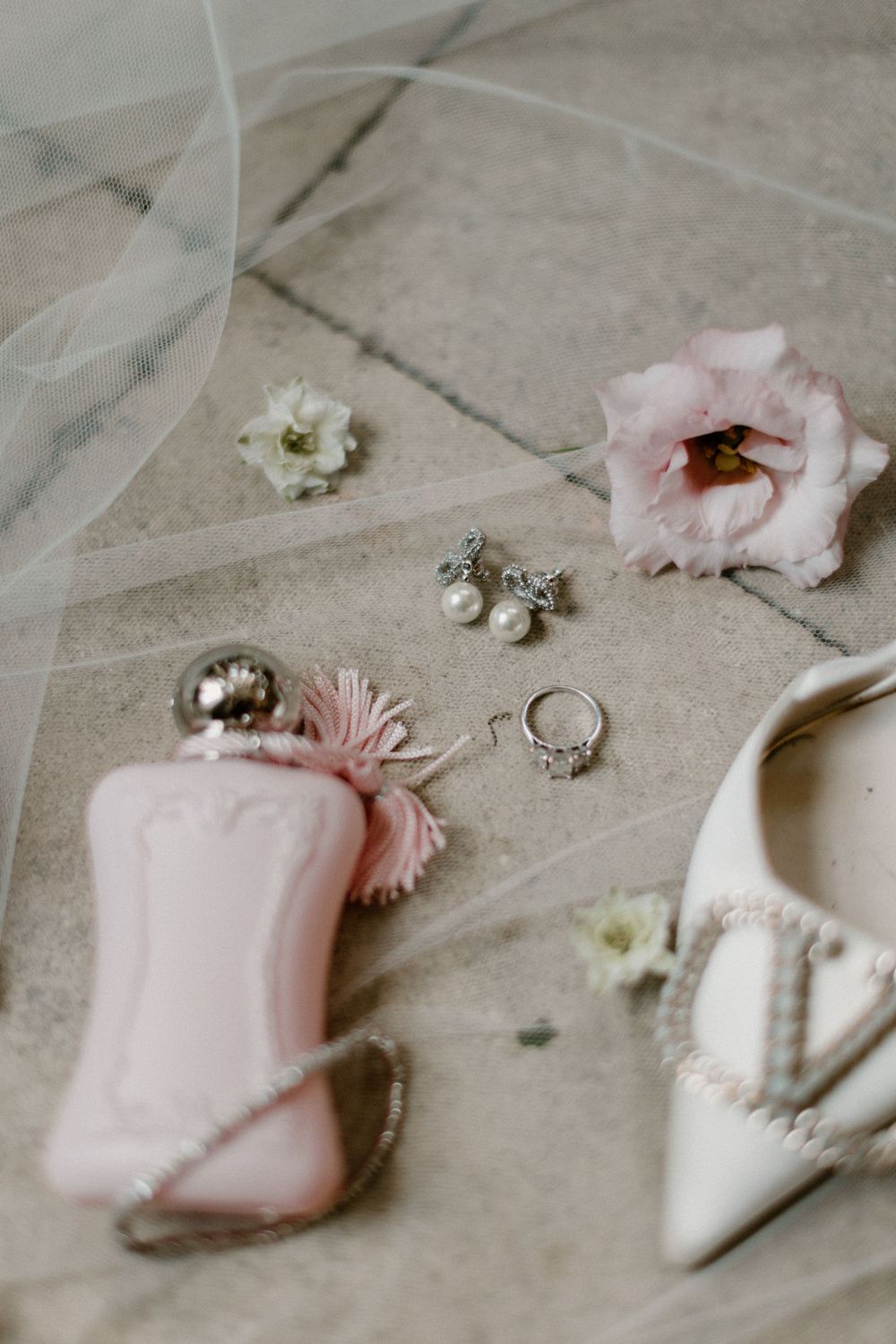 Wedding details with pink perfume bottle, jewelry, and white heel on tulle background.