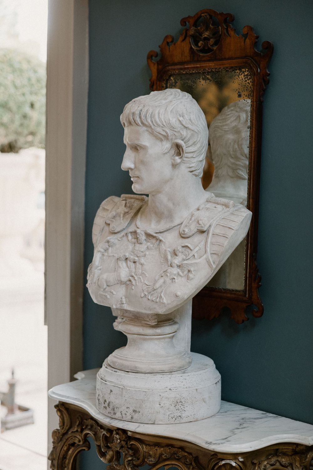 Classical marble Roman bust displayed against blue wall with ornate gold mirror.