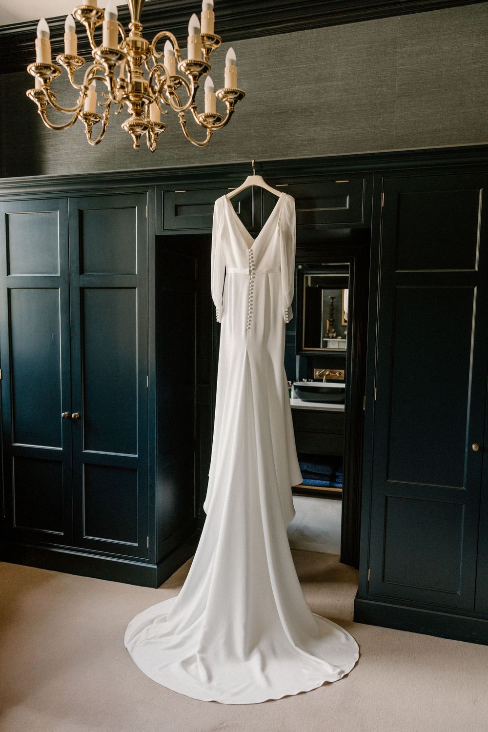 Elegant white wedding dress hanging from chandelier against dark navy paneled walls.