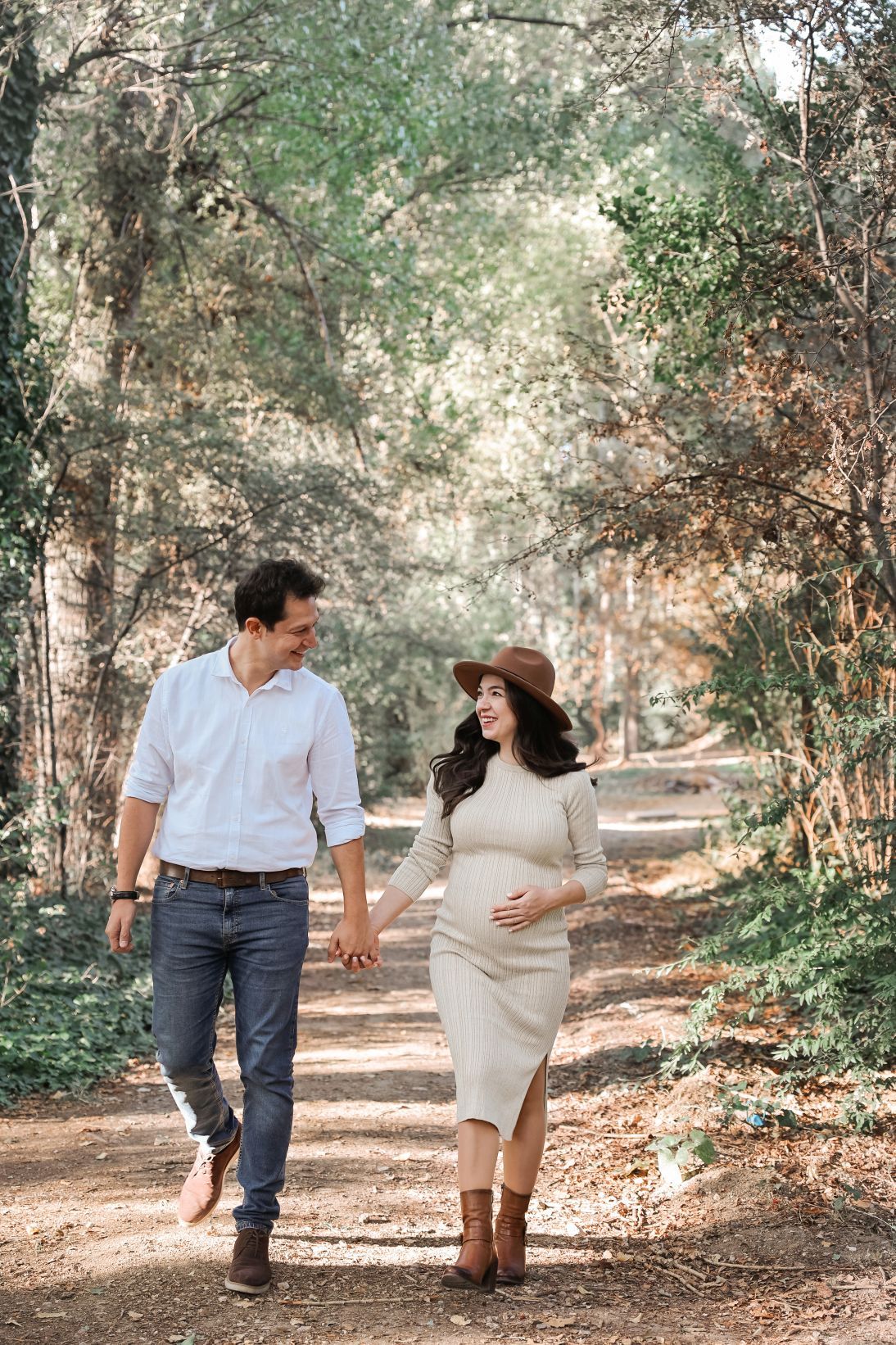 A couple walks hand in hand on a wooded path during autumn, with sunlight filtering through trees.