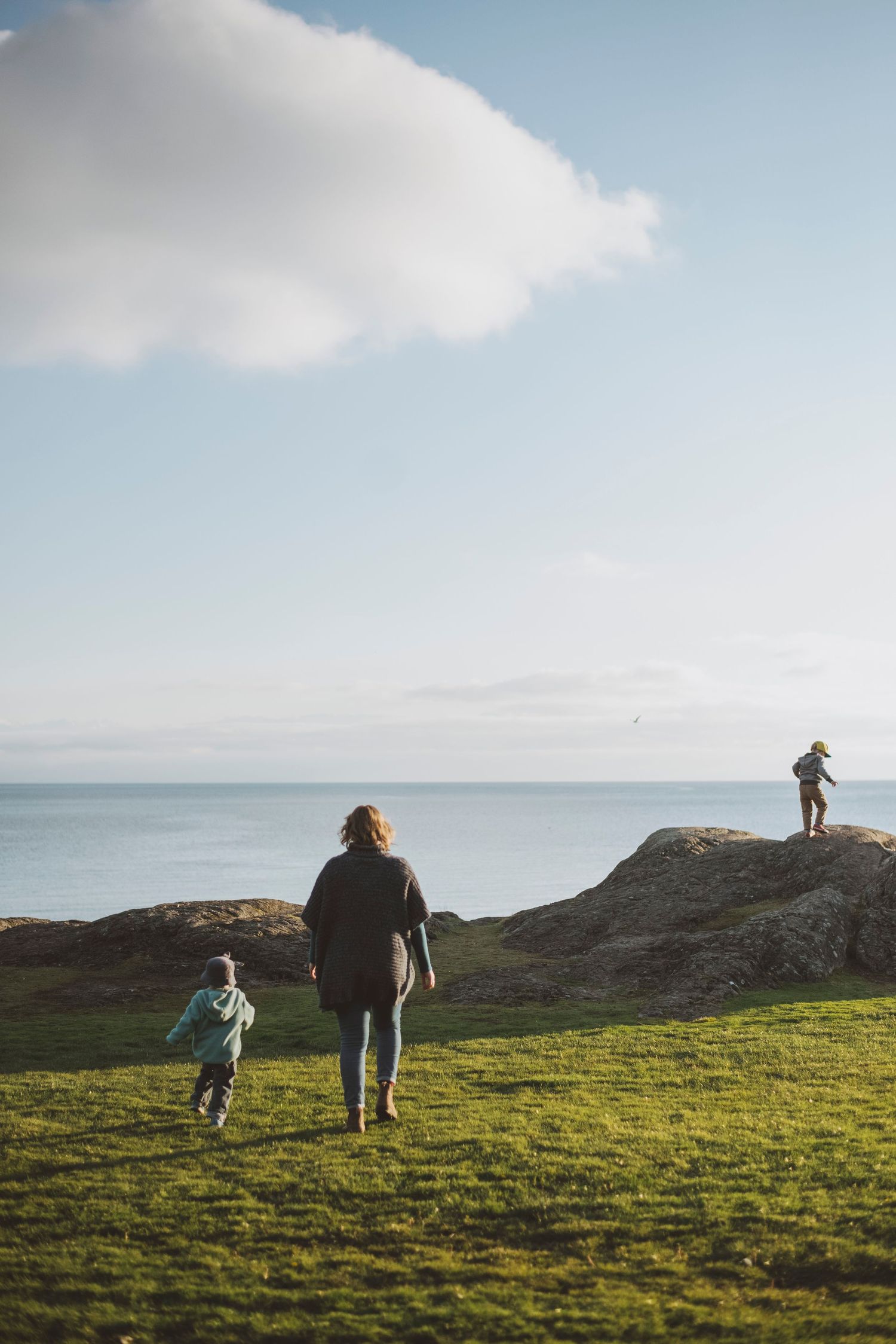 Mother and two children exploring a grassy hilltop by the ocean under a wide, open sky.