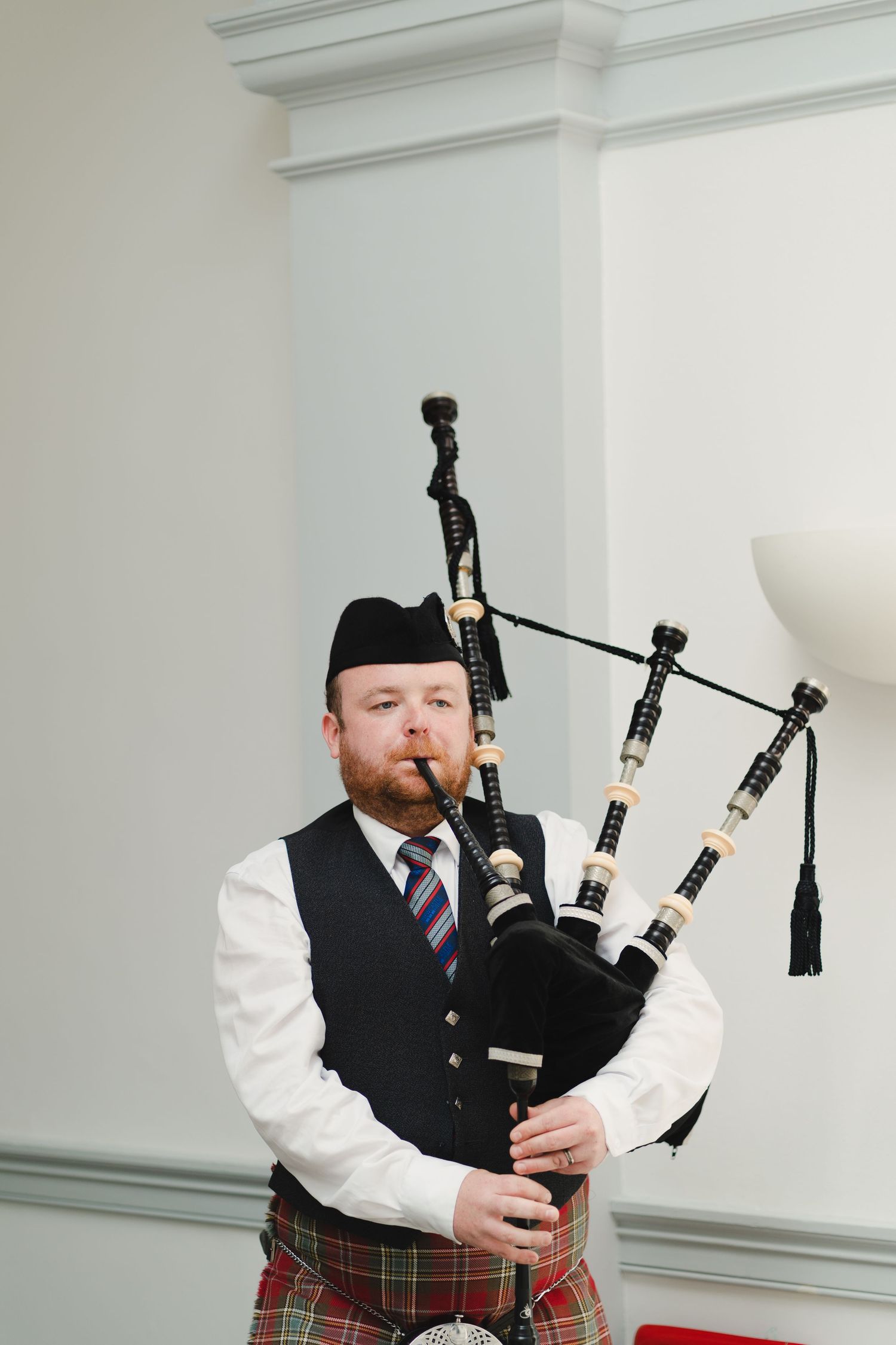 A bagpiper in traditional Scottish attire performs indoors against a white wall.