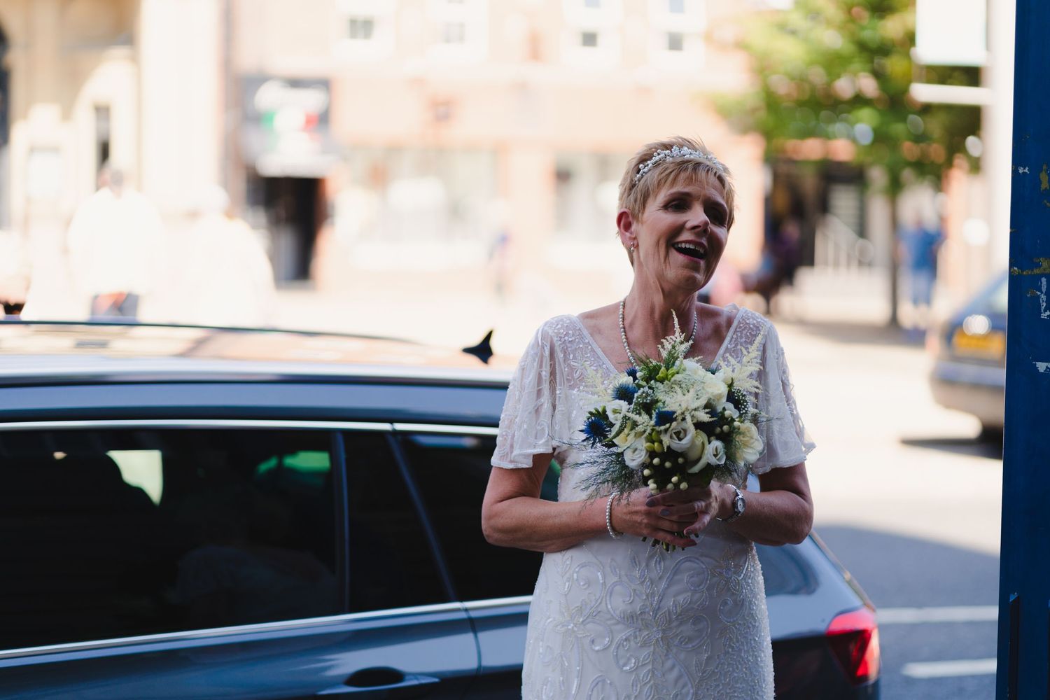 Someone in a white dress holds a floral bouquet next to a car on a sunny street.