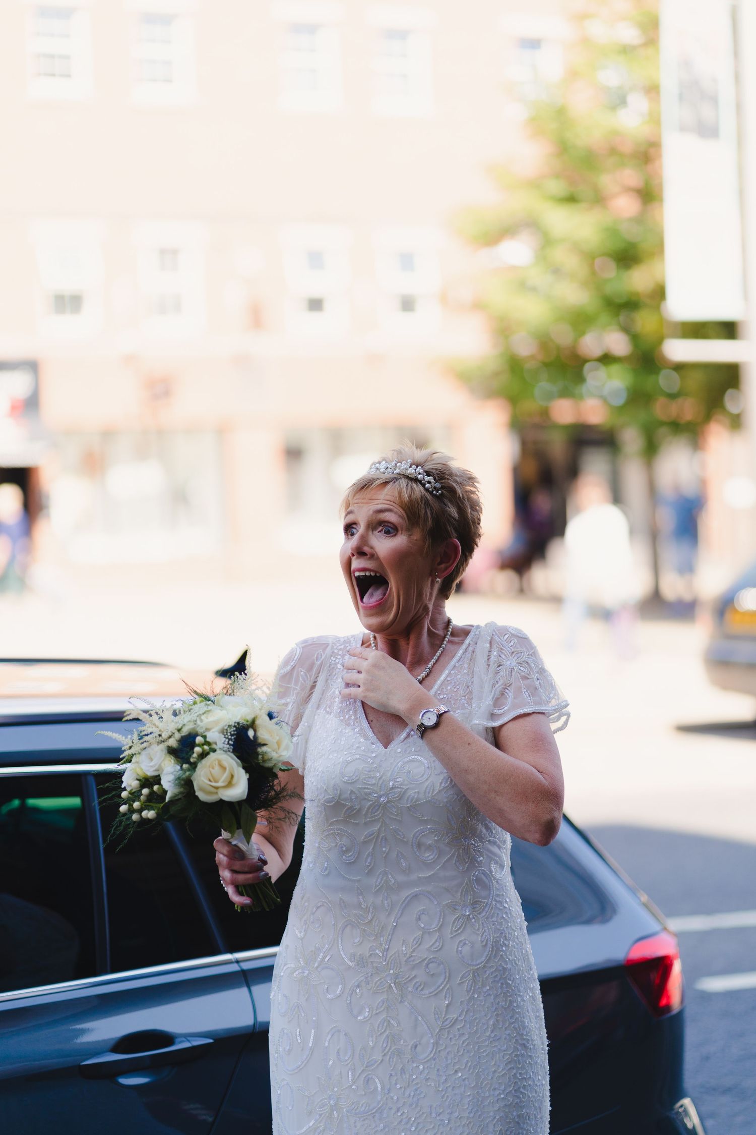 An excited person in a white lace dress holding a bouquet expresses joy outdoors.