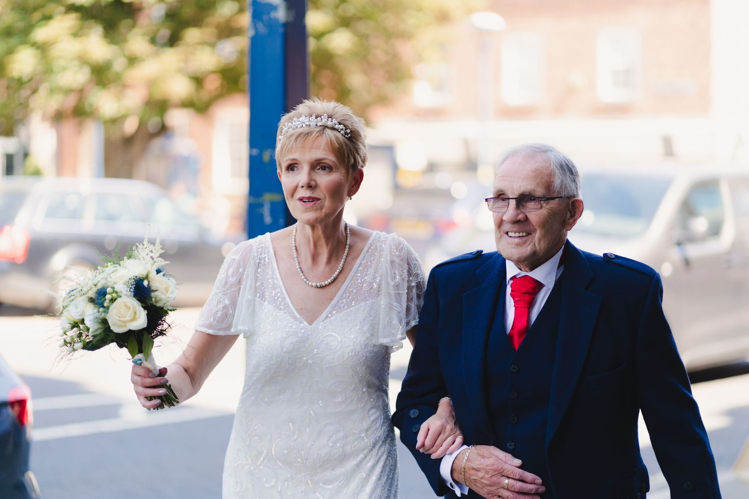 A couple walks together outdoors, with one wearing a white dress and holding a bouquet.