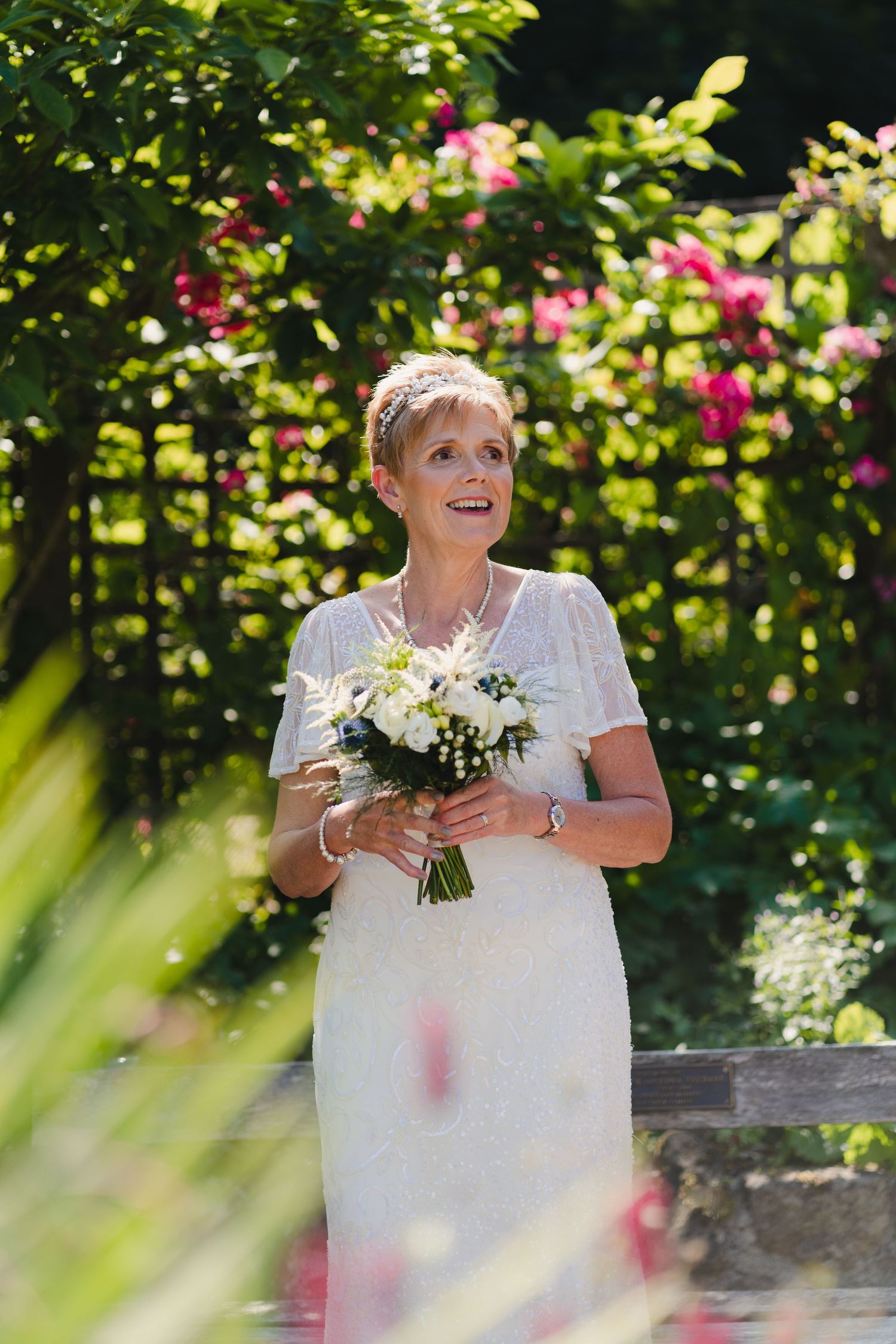 Bride holds white bouquet while standing in sunny garden with pink flowers.