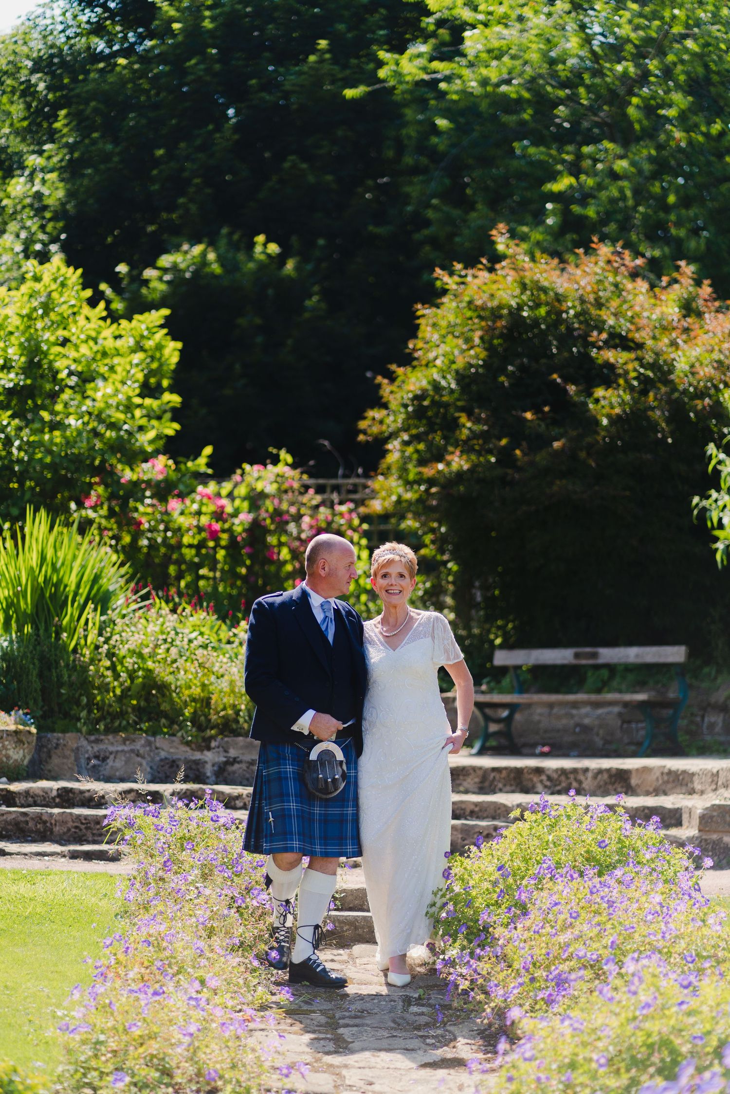 Couple walks garden path with groom in traditional Scottish kilt and bride in elegant white dress.