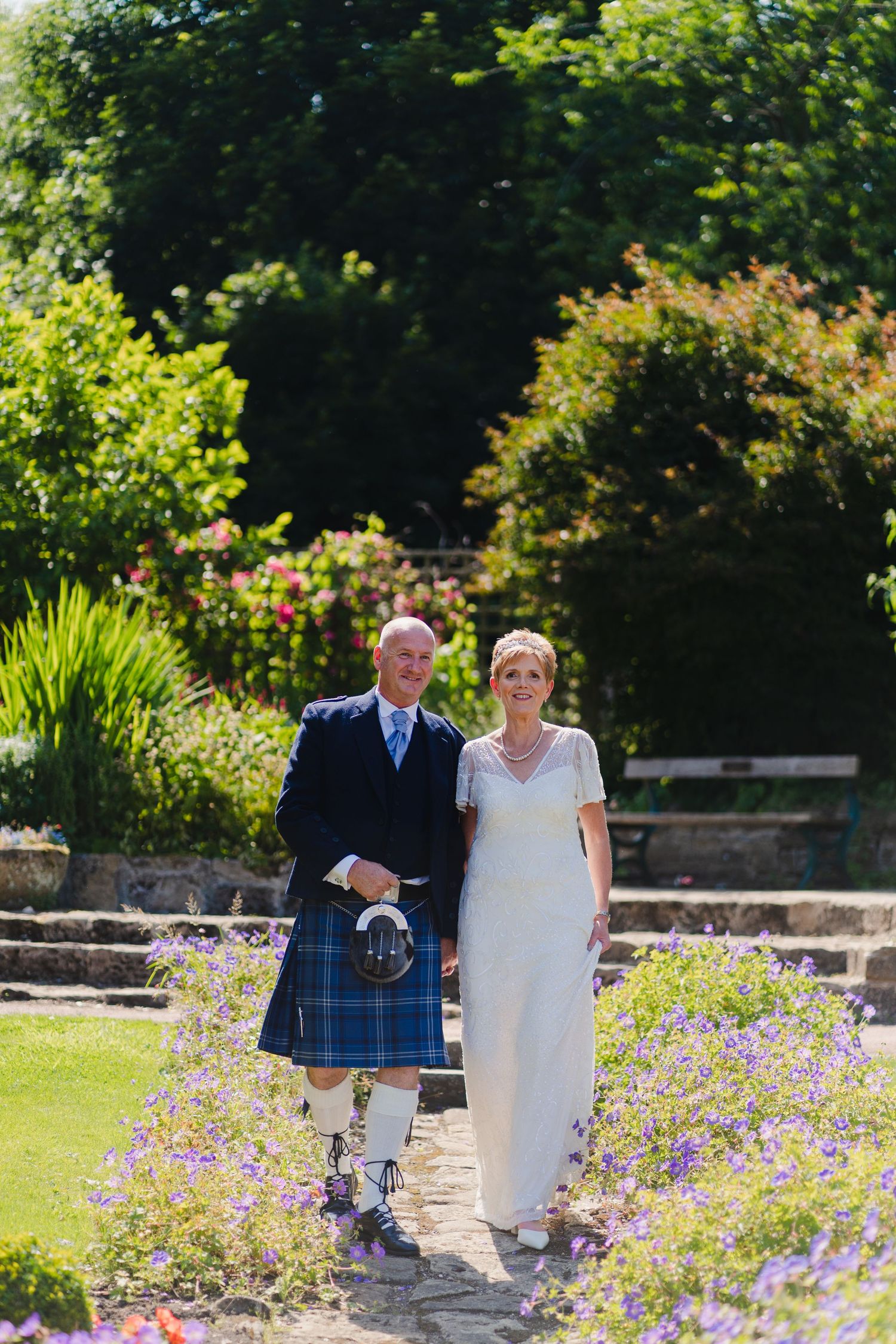 Sunlit garden wedding portrait with traditional Scottish attire and blooming flowers.