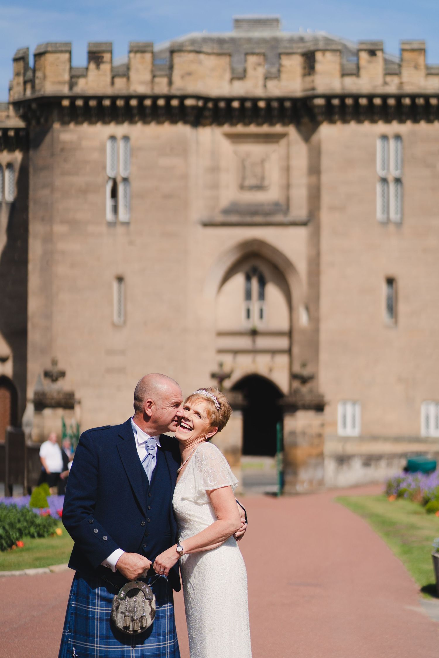 Happy couple poses outside historic castle entrance in Scottish wedding attire.