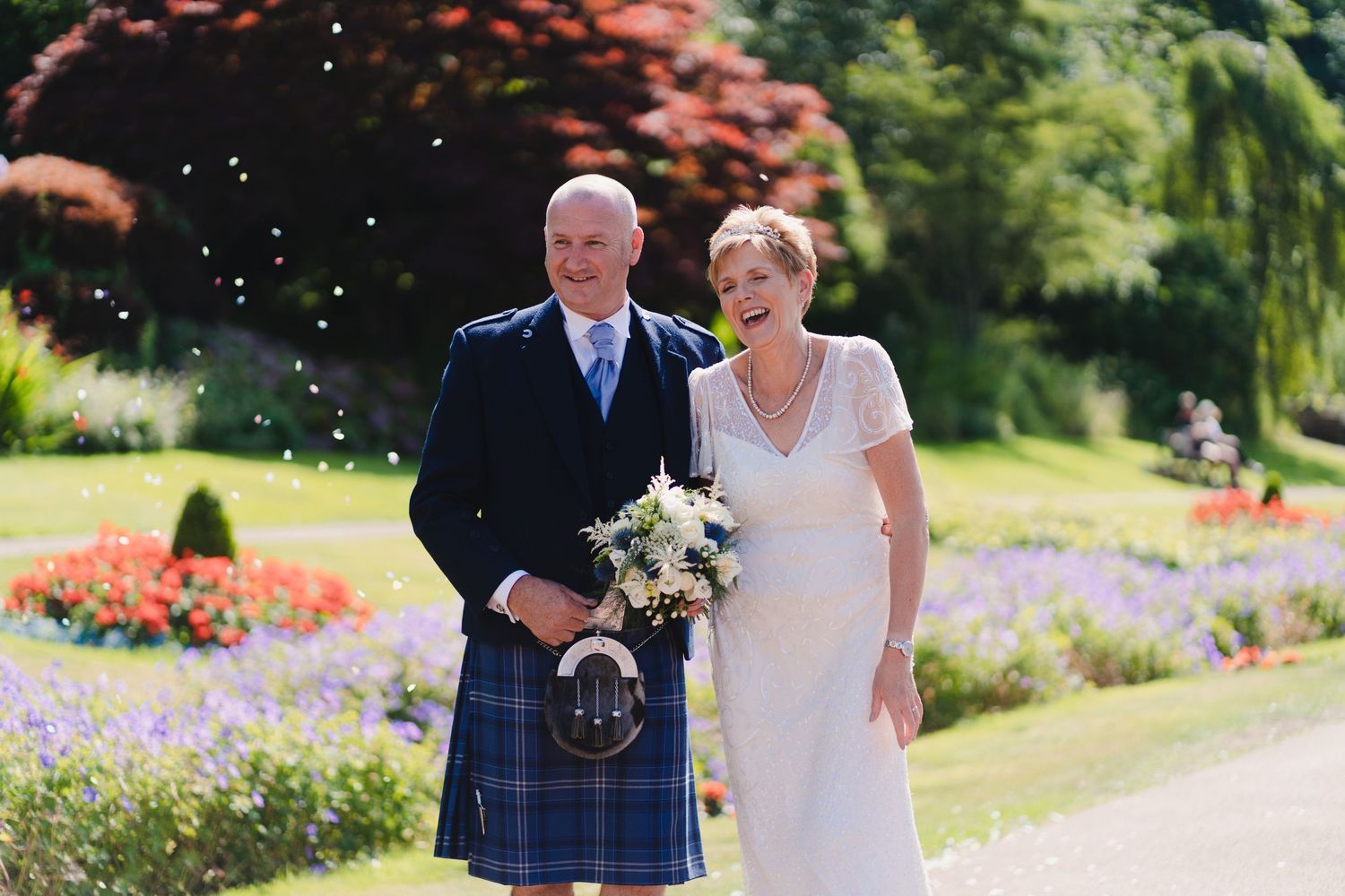 A couple poses in a garden on their wedding day, with the groom wearing a traditional Scottish kilt.