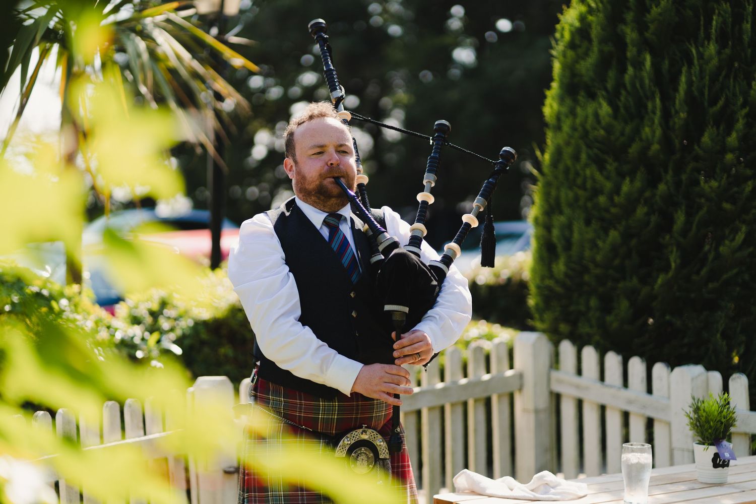 A bagpiper performs outdoors in traditional Scottish attire near a white picket fence.