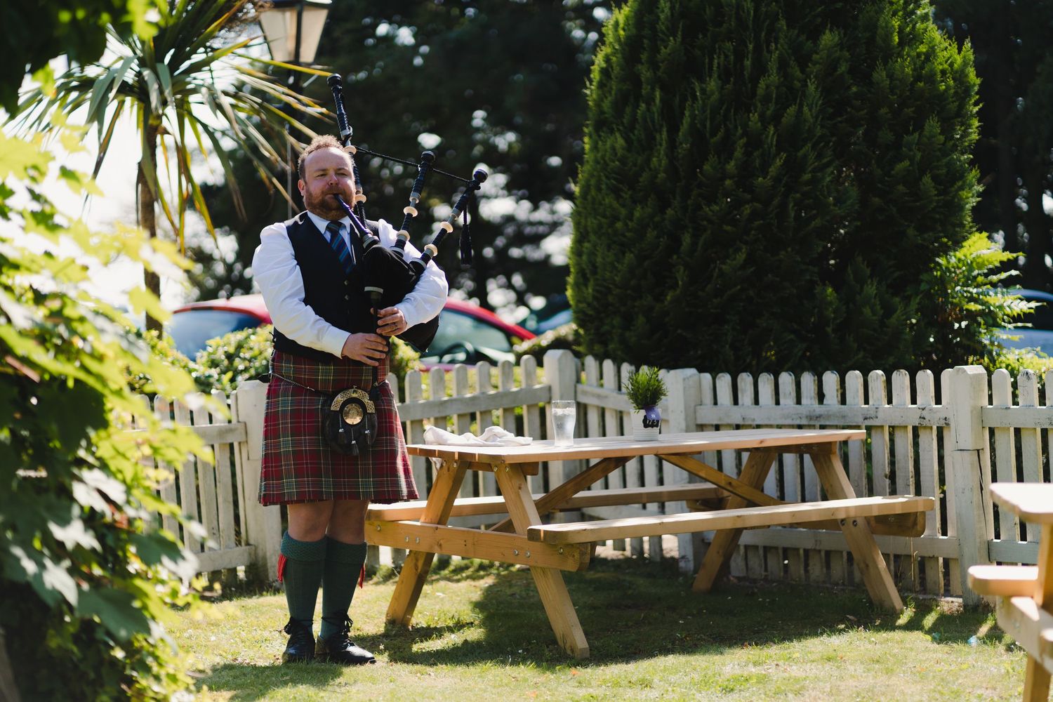 A kilted musician plays bagpipes in a sunny garden setting with wooden picnic tables.