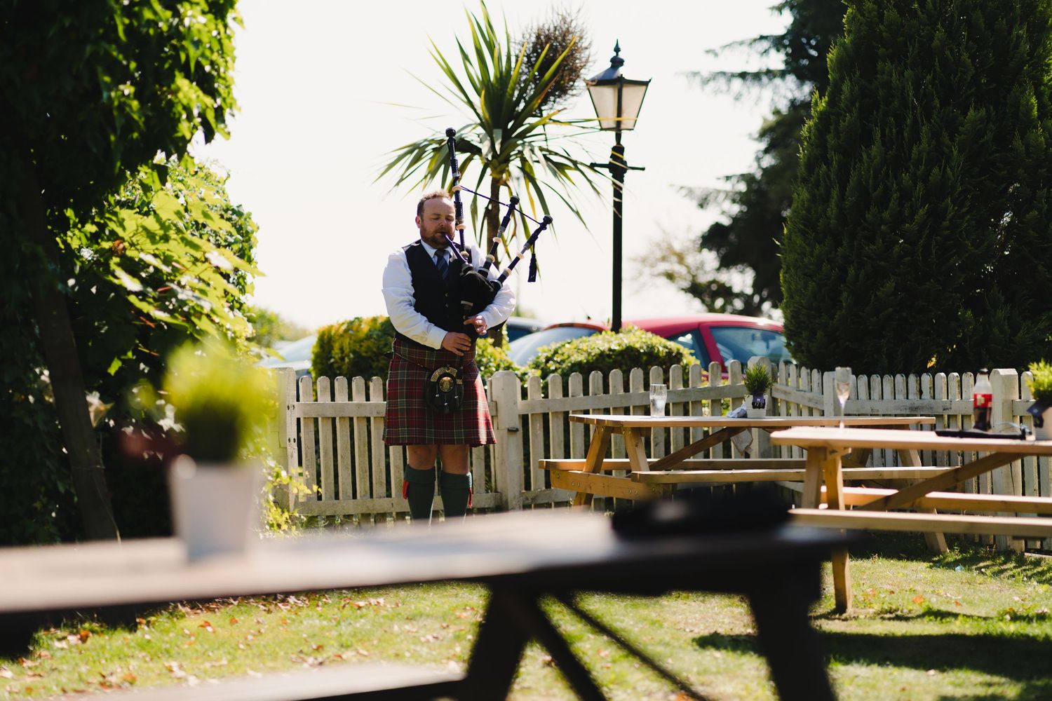 A bagpiper performs in a garden setting with picnic tables and a vintage lamppost.