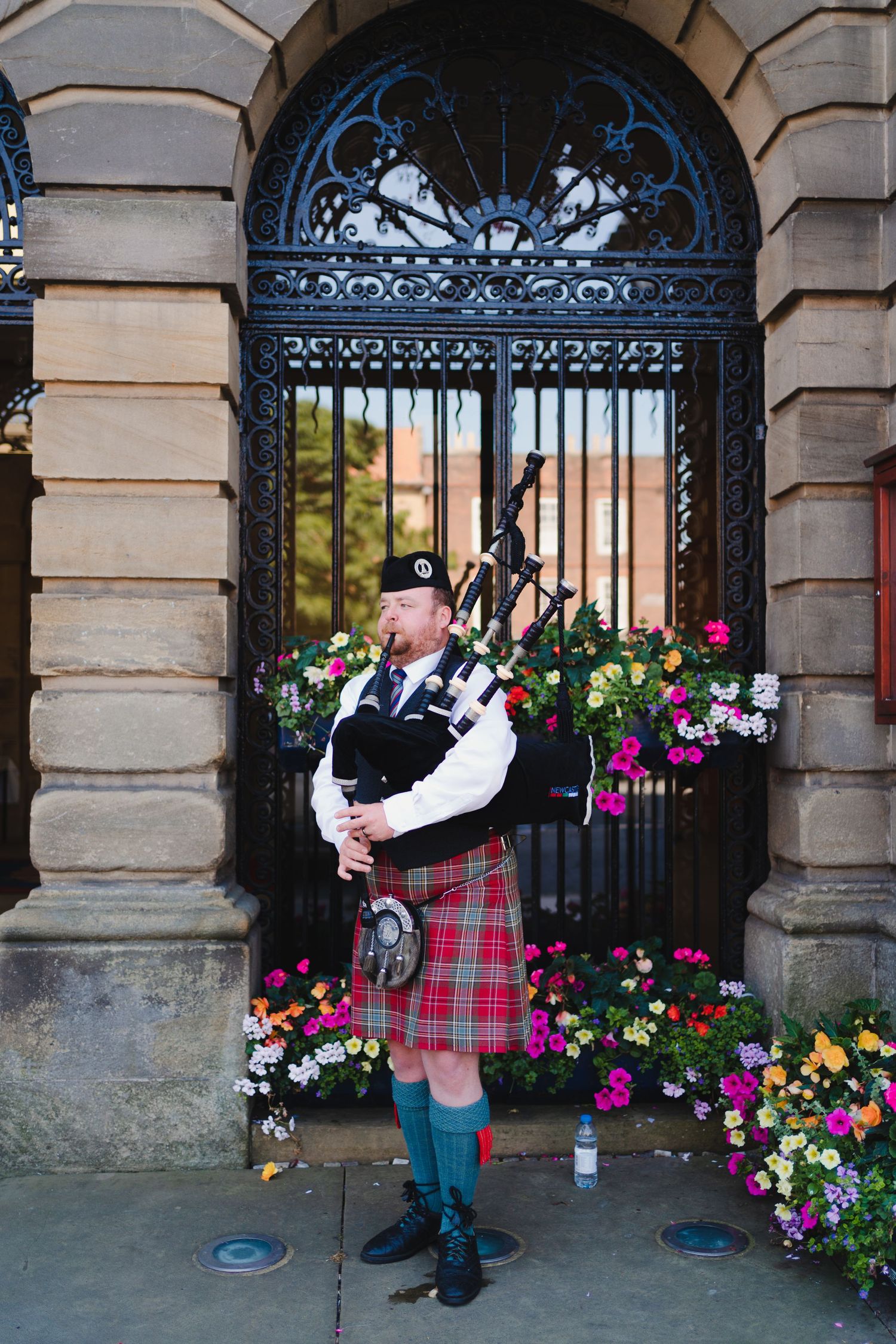 A bagpiper in traditional Scottish dress performs by an ornate iron gate with colorful flowers.