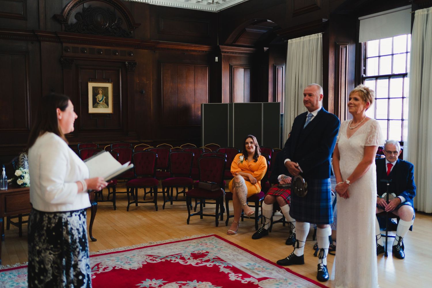 A wedding ceremony takes place in a wood-paneled room with red carpet and chairs.