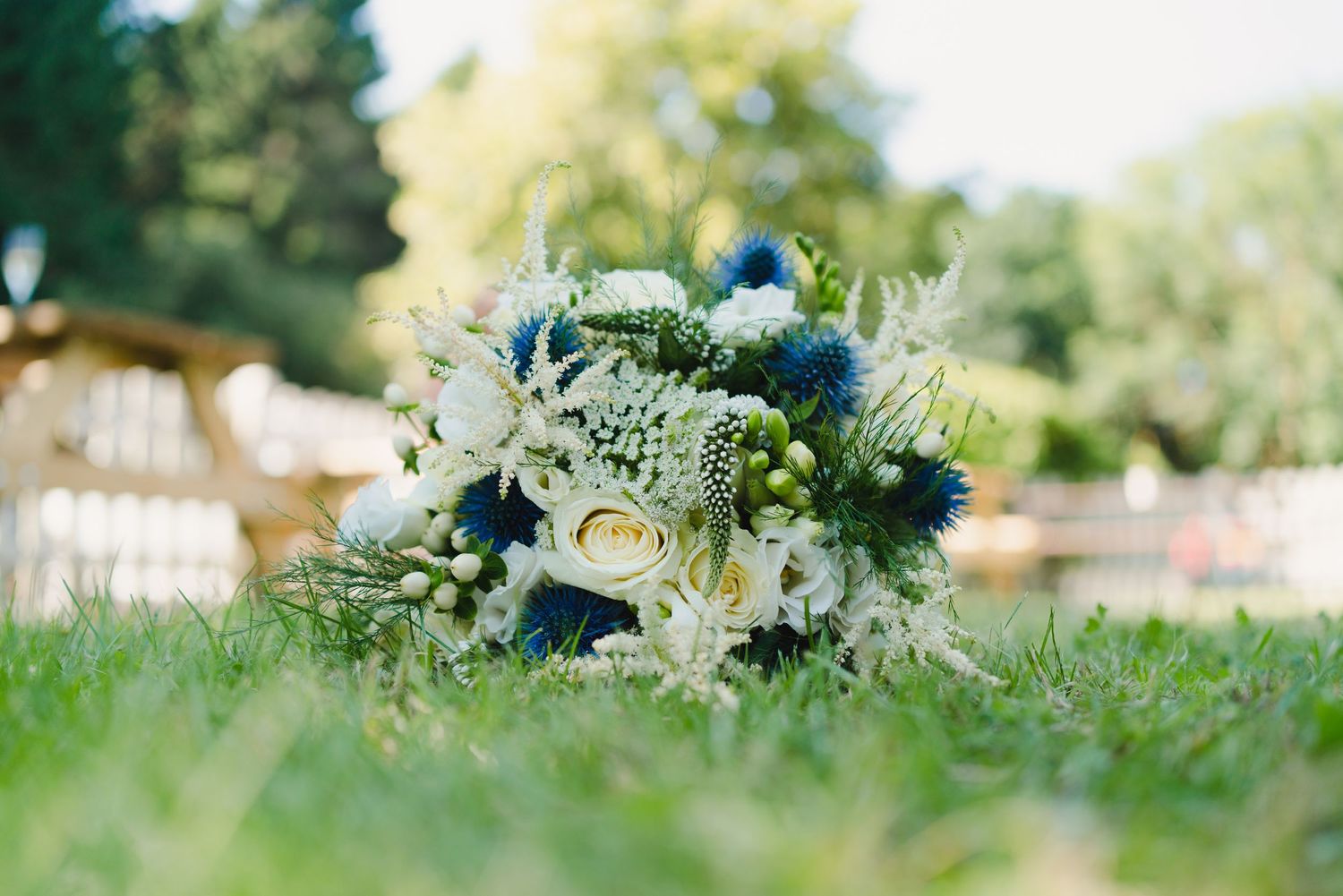 A wedding bouquet with blue and white flowers lies on green grass in a garden setting.