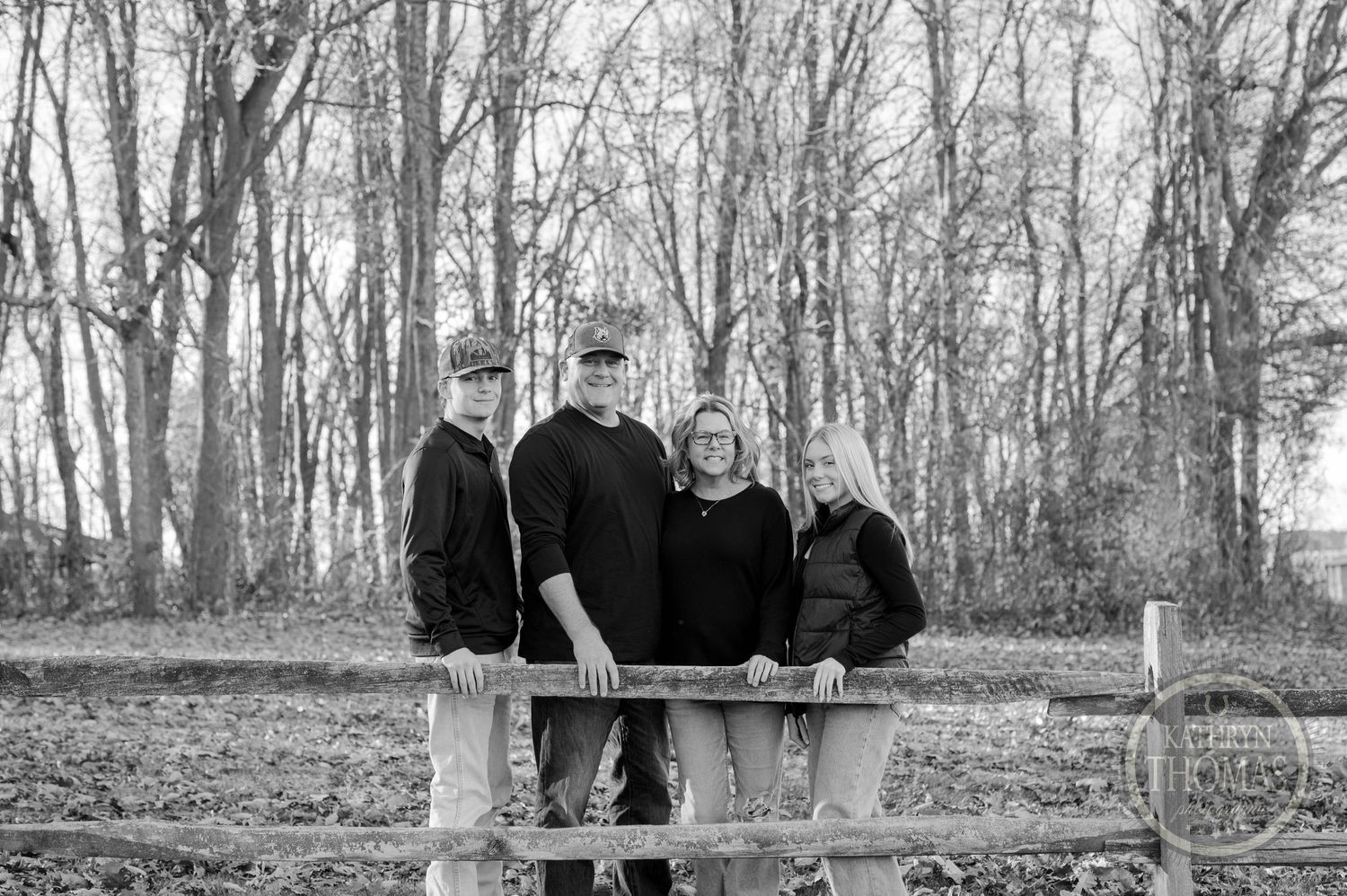 Black and white portrait of group standing at wooden fence with bare trees in background.